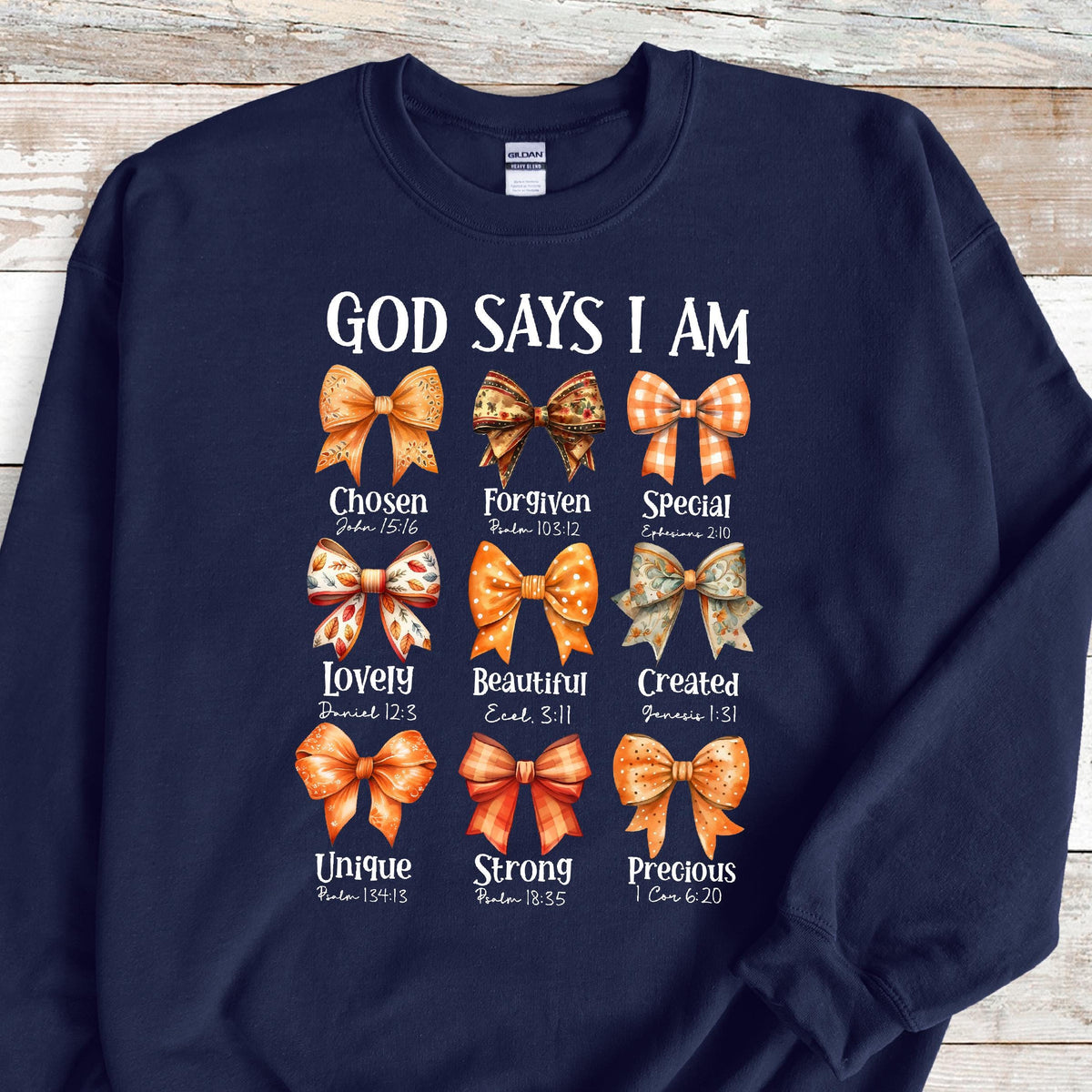 May include: A navy blue crewneck sweatshirt featuring the white text GOD SAYS I AM above nine distinct orange and brown patterned bows. Each bow is paired with a descriptive word and a Bible verse reference, including Chosen, Forgiven, Special, Lovely, Beautiful, Created, Unique, Strong, and Precious.