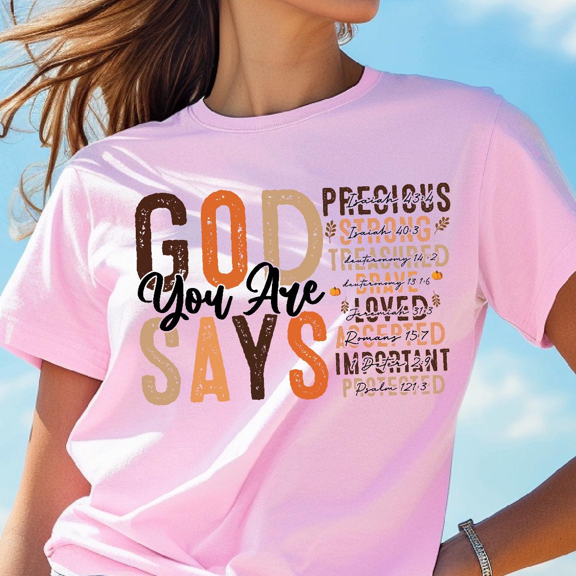 May include: Pink t-shirt with the words "GOD You Are SAYS" in a mix of brown, orange, and beige. The shirt also features the words "PRECIOUS, STRONG, TREASURED, BRAVE, LOVED, ACCEPTED, IMPORTANT" with Bible verses.