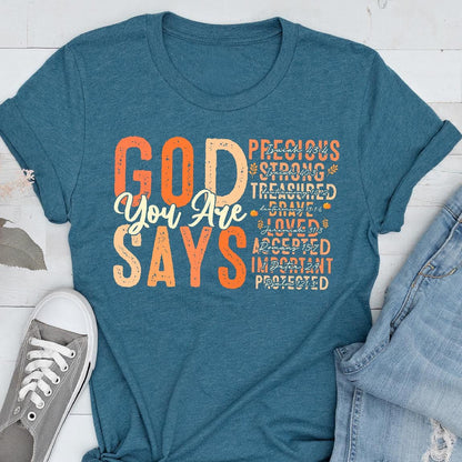 May include: Teal t-shirt with the words "GOD You Are SAYS" in orange and white. Additional words like "Precious", "Strong", and "Loved" are also printed on the shirt. The shirt is paired with blue jeans and a gray sneaker.