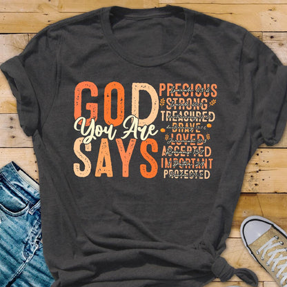 May include: Dark gray t-shirt with the words "GOD You Are SAYS" in orange and cream lettering. Additional words like "Precious" and "Loved" are also printed on the shirt. The shirt is displayed on a wooden surface.