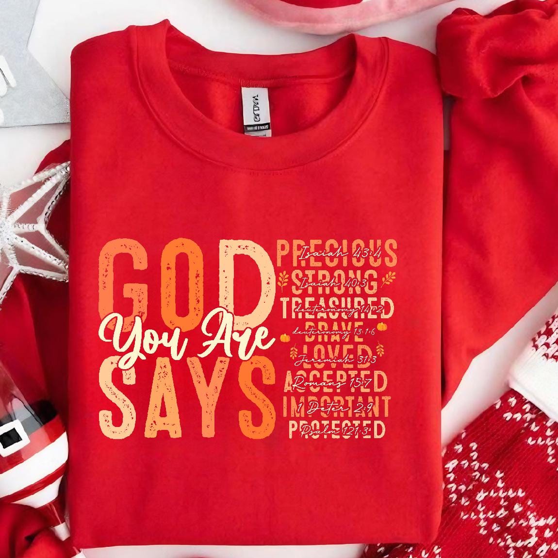 May include: Red sweatshirt with the text "GOD YOU ARE SAYS" in a gradient of orange and yellow. The text is surrounded by a list of words and bible verses, including "Precious", "Strong", "Treasured", "Brave", "Loved", "Accepted", "Important", and "Protected".