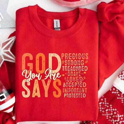 May include: Red sweatshirt with the text "GOD YOU ARE SAYS" in a gradient of orange and yellow. The text is surrounded by a list of words and bible verses, including "Precious", "Strong", "Treasured", "Brave", "Loved", "Accepted", "Important", and "Protected".