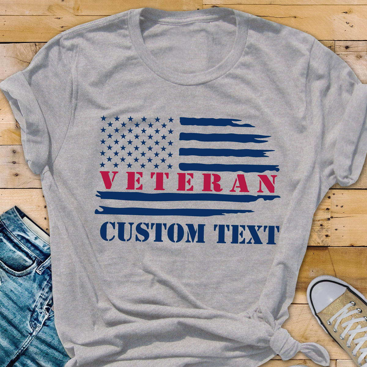 May include: A gray t-shirt featuring a distressed American flag design. The word "VETERAN" is in red, and "CUSTOM TEXT" is in blue. The shirt is made of a soft material.