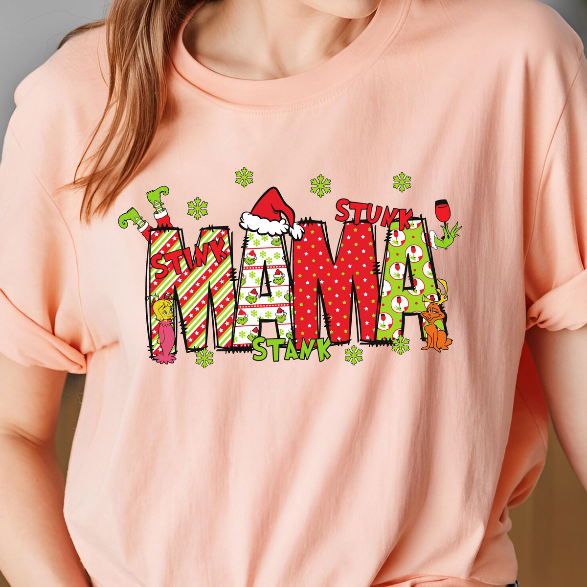 May include: A peach-colored t-shirt featuring a graphic with the text STINK MAMA STANK. The letters are filled with red and green patterns, including stripes, polka dots, and small character illustrations, accented by green snowflakes.