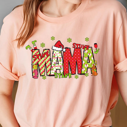 May include: A peach-colored t-shirt featuring a graphic with the text STINK MAMA STANK. The letters are filled with red and green patterns, including stripes, polka dots, and small character illustrations, accented by green snowflakes.