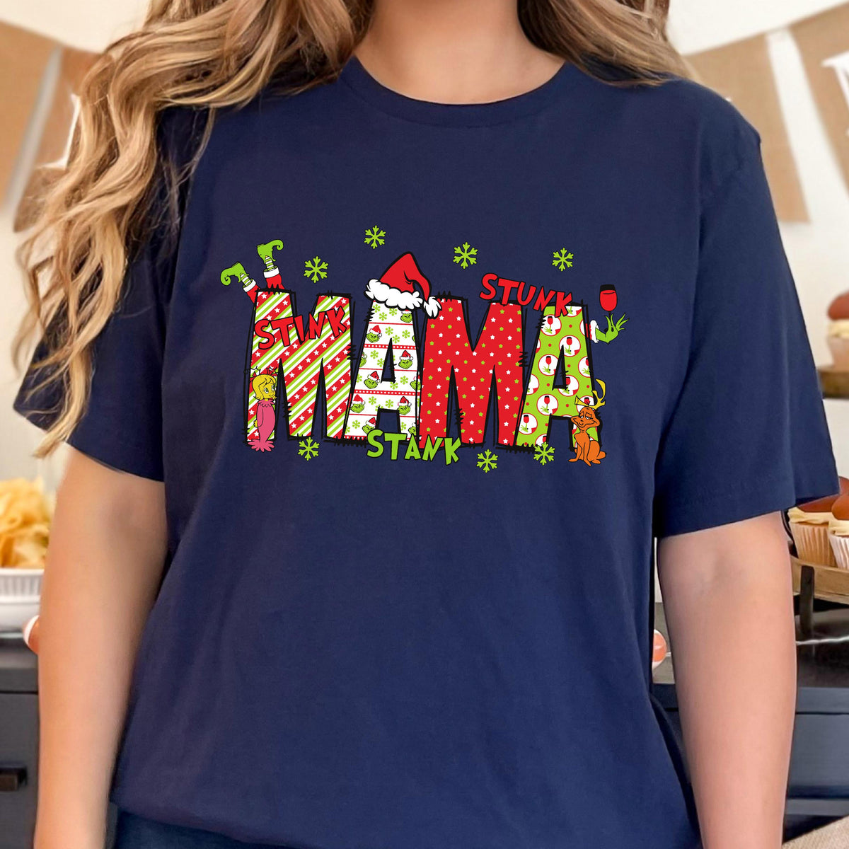 May include: A navy blue short-sleeve t-shirt featuring the word MAMA in large, patterned letters. The letters include red and green stripes, polka dots, and Grinch-themed graphics. A Santa hat sits on the letter A, and green snowflakes surround the text.