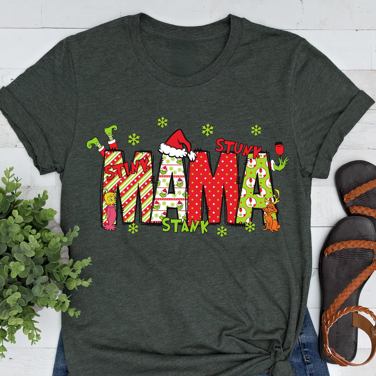 May include: A dark charcoal gray short-sleeve t-shirt featuring the word MAMA in large, patterned block letters. The letters are decorated with red and green stripes, polka dots, and Grinch-themed graphics, including a Santa hat and snowflakes.