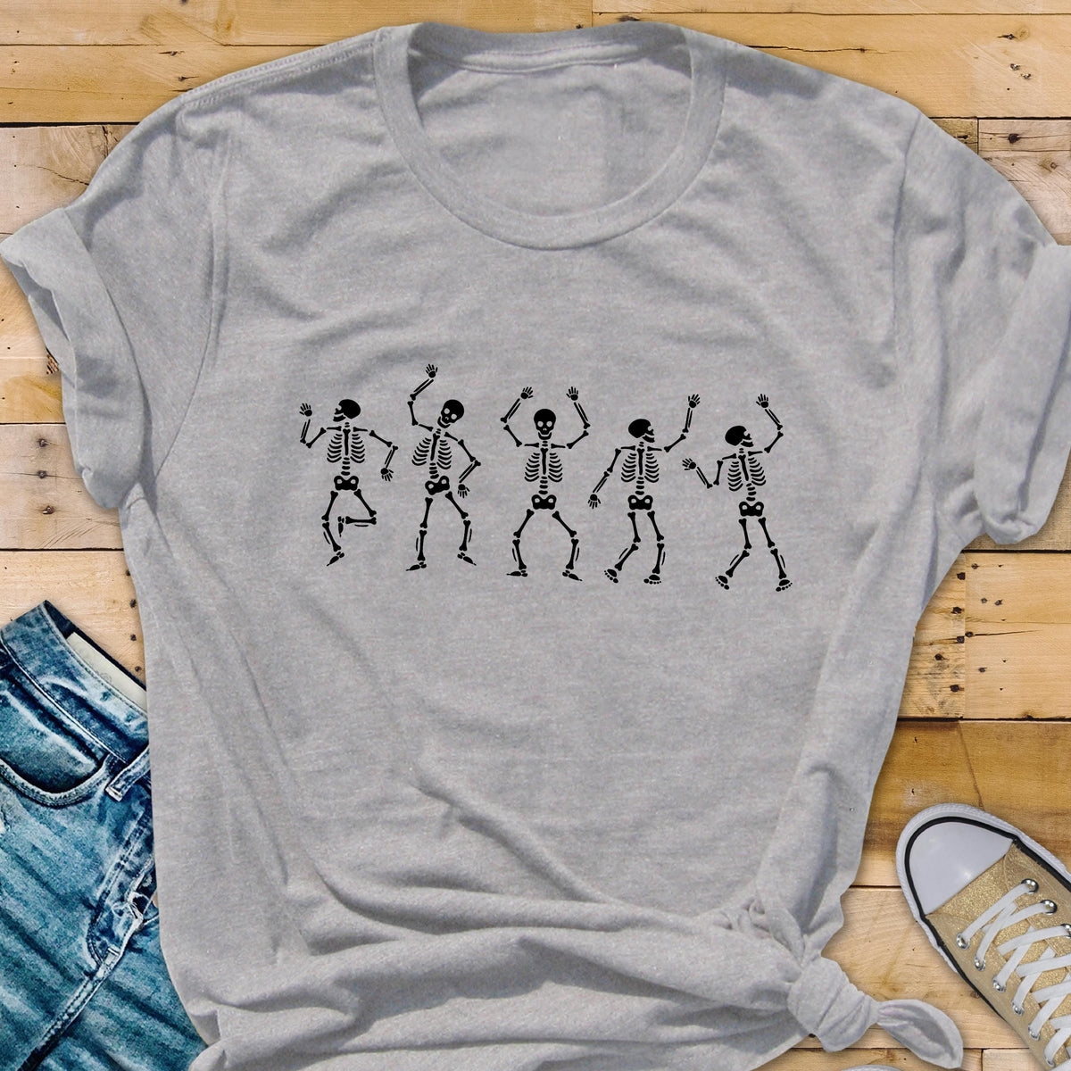 May include: A light gray t-shirt featuring a black graphic of five dancing skeletons. The skeletons are in various poses with their arms raised. The shirt is made of a soft material and has short sleeves. The shirt is a casual wear item.
