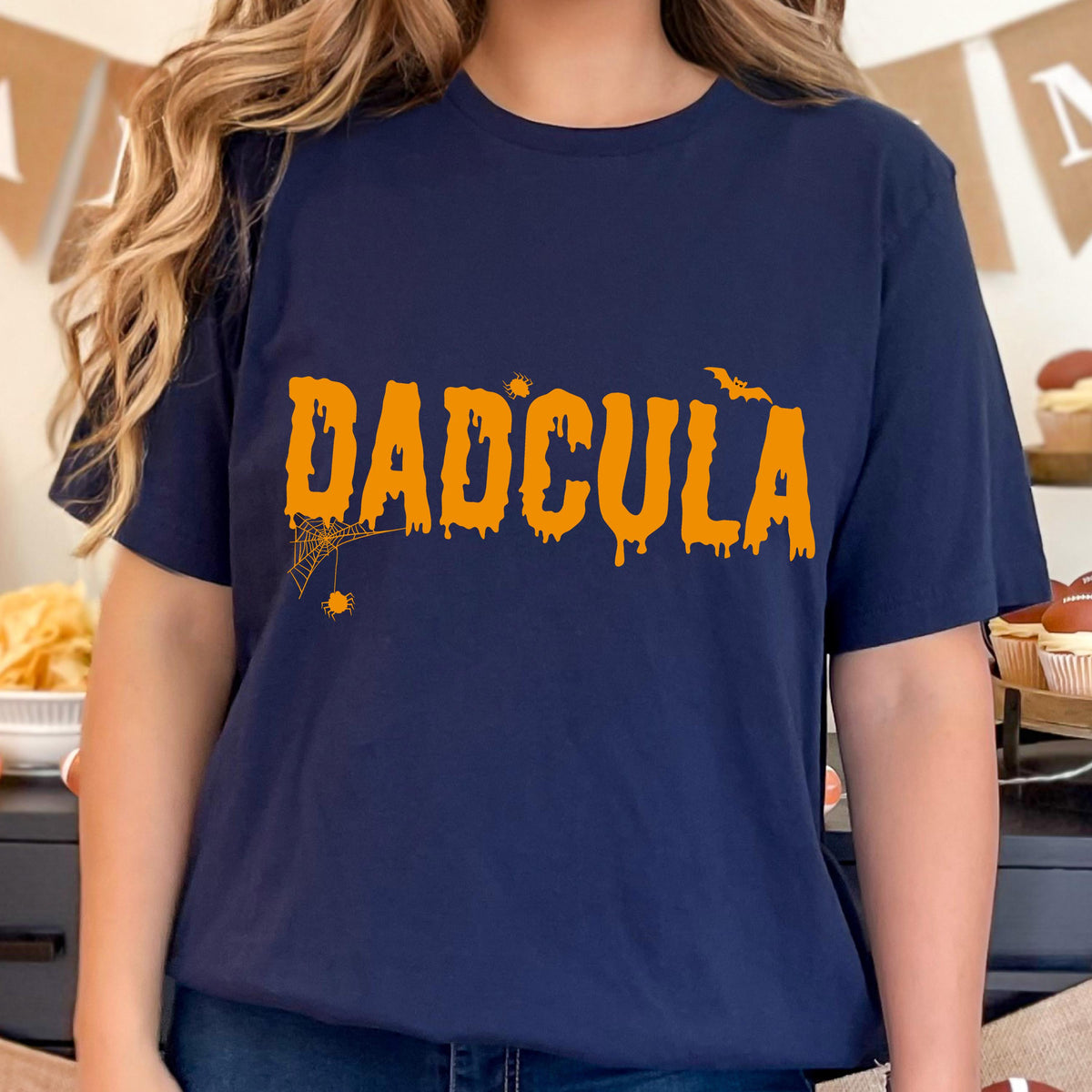 May include: A navy blue short-sleeve t-shirt featuring the orange text DADCULA in a dripping font. The design includes a small spider web with a spider hanging from the letter D, a small spider above the letter C, and a flying bat above the letter L.