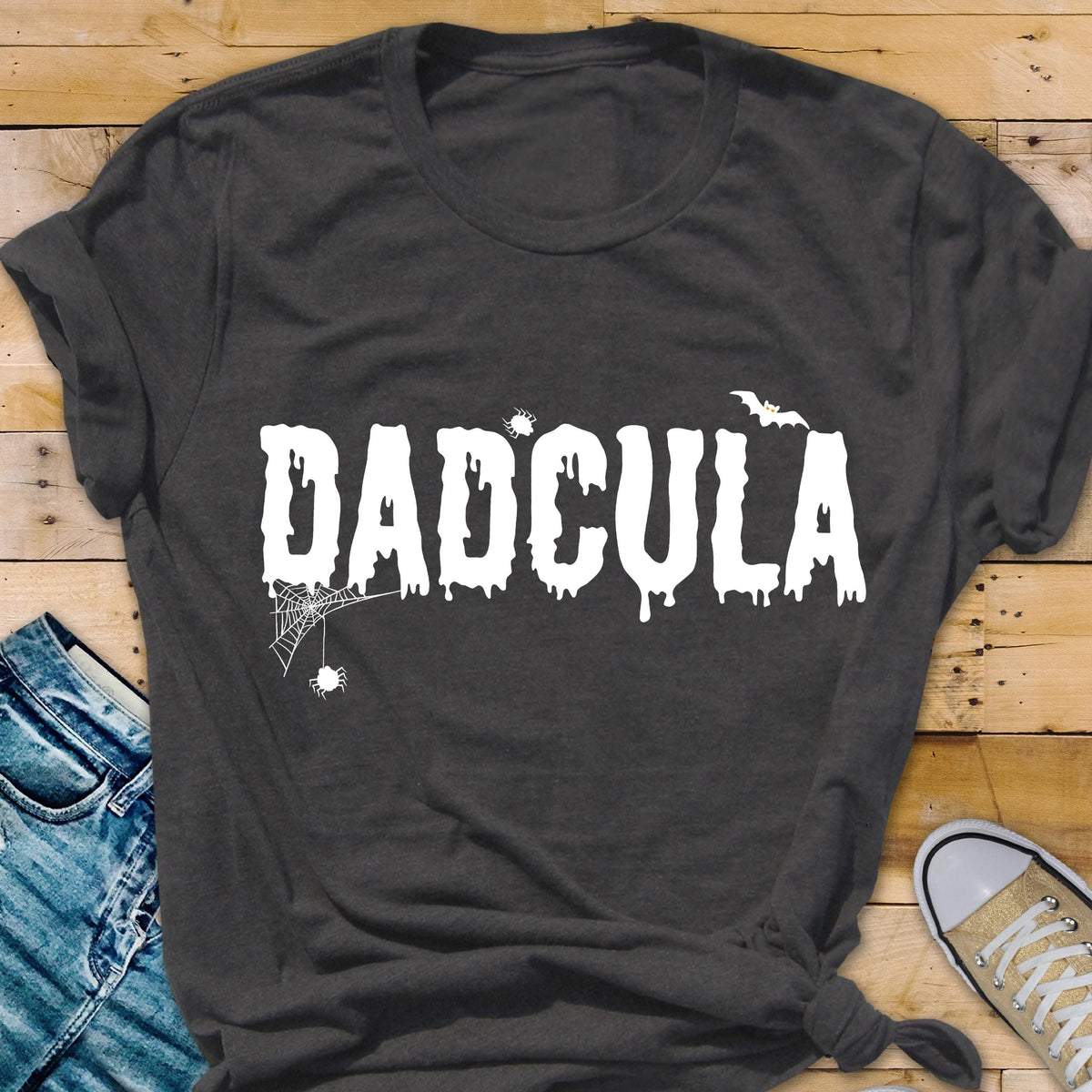 May include: A dark gray short-sleeved t-shirt featuring the white text DADCULA in a dripping font. The design includes a small spider hanging from a web on the left, a tiny spider above the letter C, and a small bat silhouette above the letter A.
