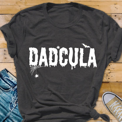 May include: A dark gray short-sleeved t-shirt featuring the white text DADCULA in a dripping font. The design includes a small spider hanging from a web on the left, a tiny spider above the letter C, and a small bat silhouette above the letter A.