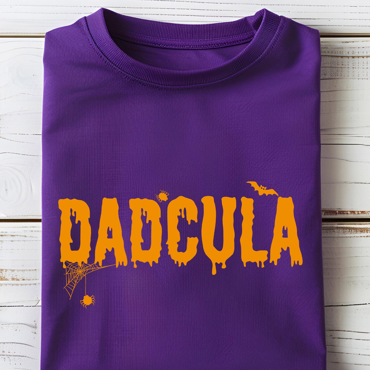 May include: A purple short-sleeved t-shirt featuring the orange text DADCULA in a dripping, horror-style font. The design includes a small spider web with a hanging spider, a tiny spider above the letter A, and a flying bat silhouette.