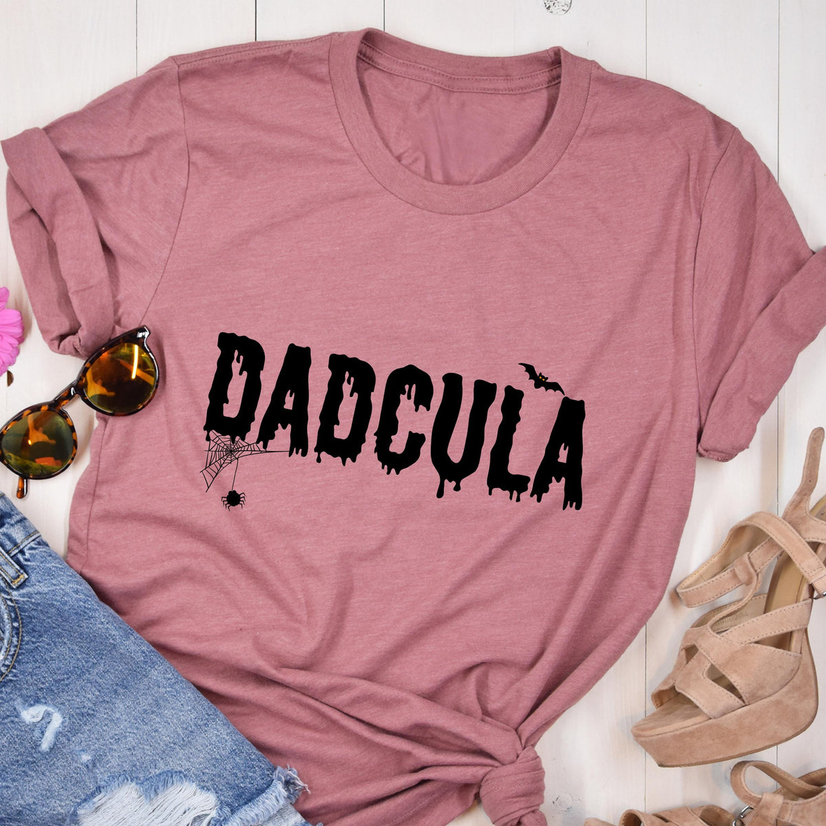 May include: A dusty rose short-sleeve t-shirt featuring the black text DADCULA in a dripping, horror-inspired font. The design includes a small black spider hanging from a web on the first letter D and a small black bat silhouette near the letter U.