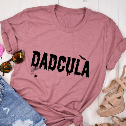 May include: A dusty rose short-sleeve t-shirt featuring the black text DADCULA in a dripping, horror-inspired font. The design includes a small black spider hanging from a web on the first letter D and a small black bat silhouette near the letter U.