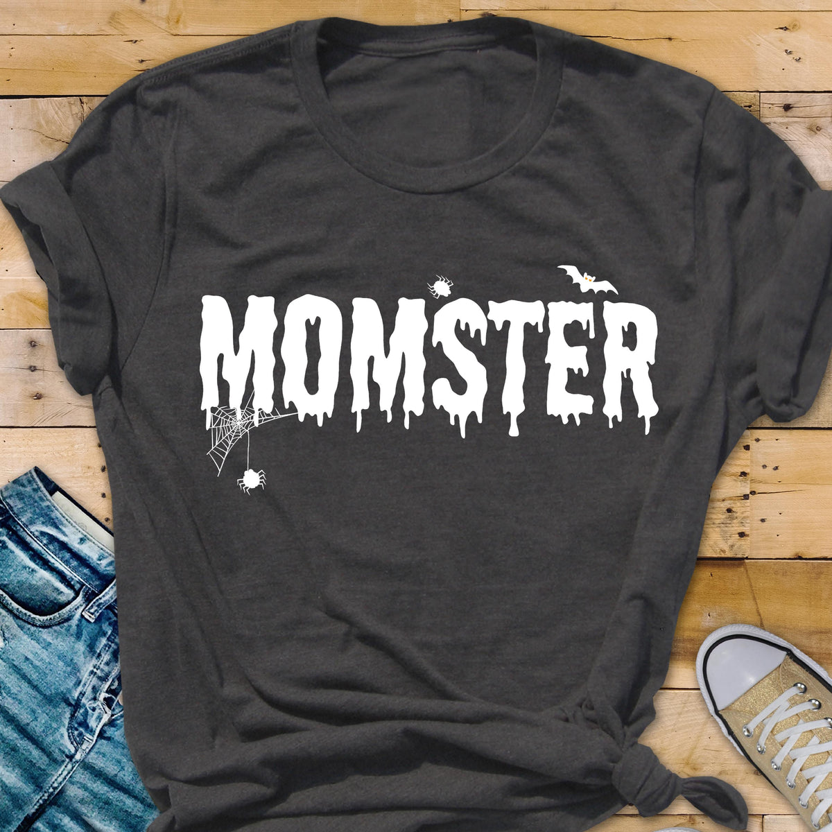 May include: Dark gray t-shirt with the word "MOMSTER" in white, dripping font. A spider web and spider are on the left, and a bat is above the "R". The shirt is folded at the bottom and has a casual style.