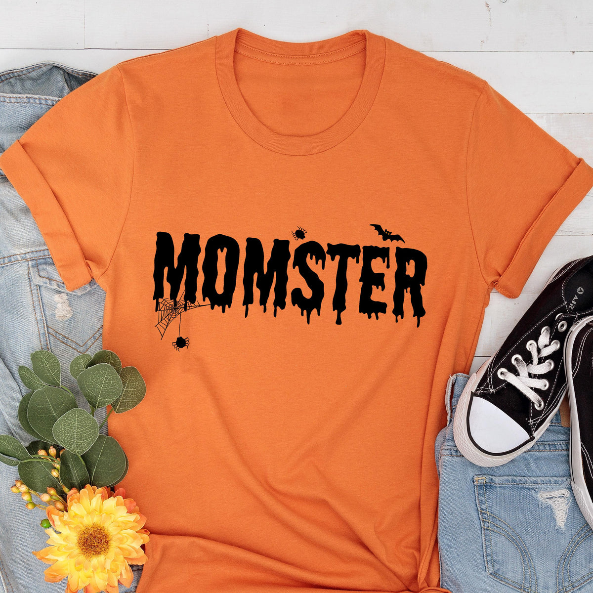 May include: Orange t-shirt with the word "MOMSTER" in black, dripping font. A spider web, spider, and bat are above the word. The shirt is folded at the sleeves. A pair of black sneakers and blue jeans are in the background.