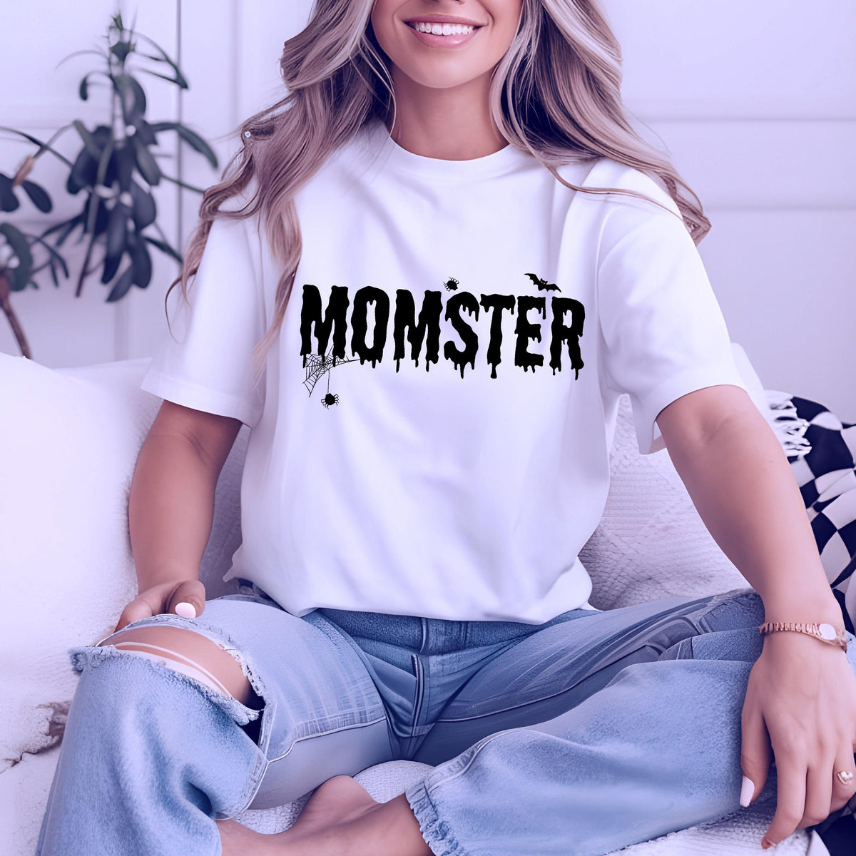 May include: White t-shirt with the word "MOMSTER" in black, dripping font, with a spider web and bat graphic. The shirt is short-sleeved and worn with light blue jeans. A fun Halloween-themed tee.