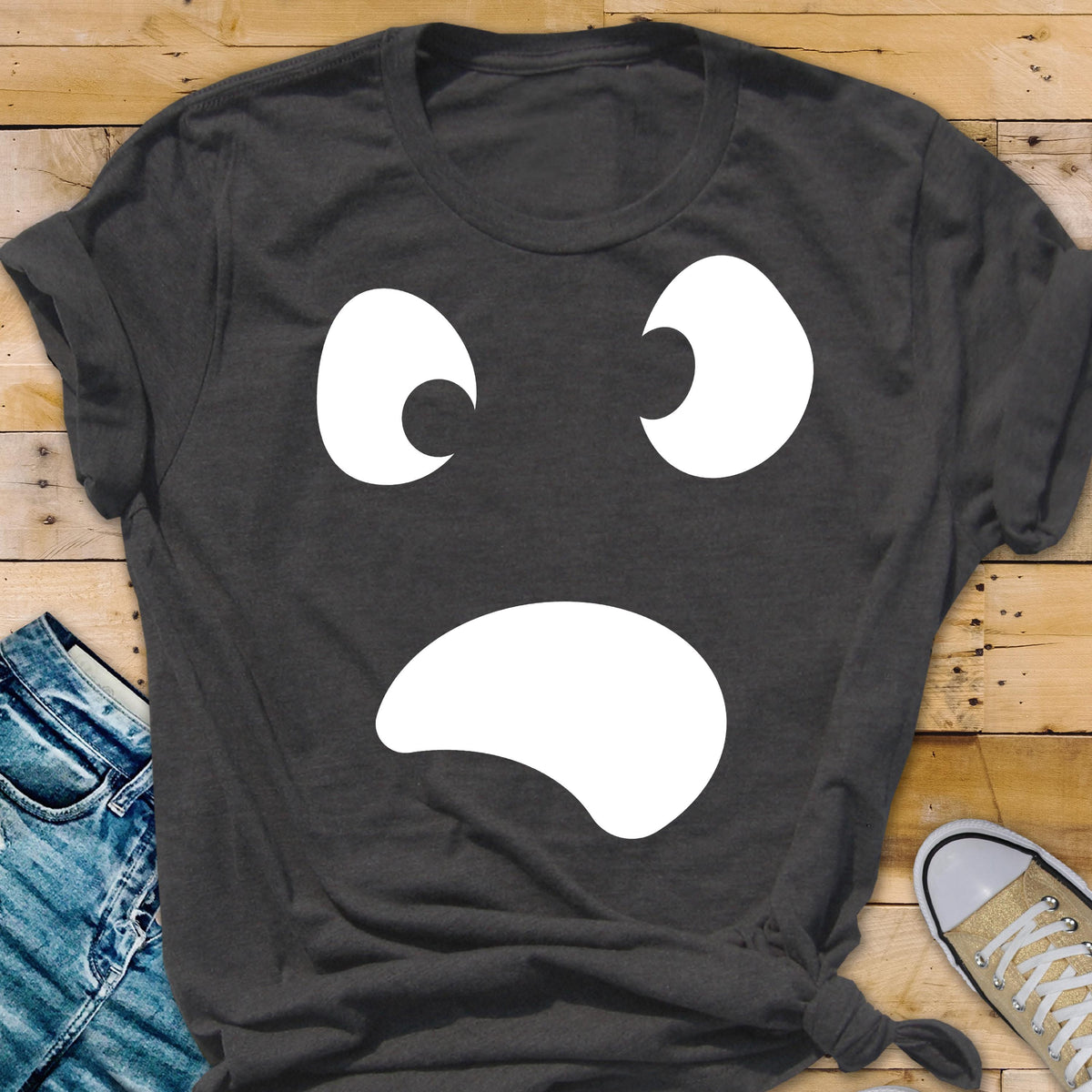 May include: Dark gray t-shirt featuring a white, cartoonish face with two crescent-shaped eyes and a curved mouth. The design is centered on the shirt, creating a simple yet expressive graphic.