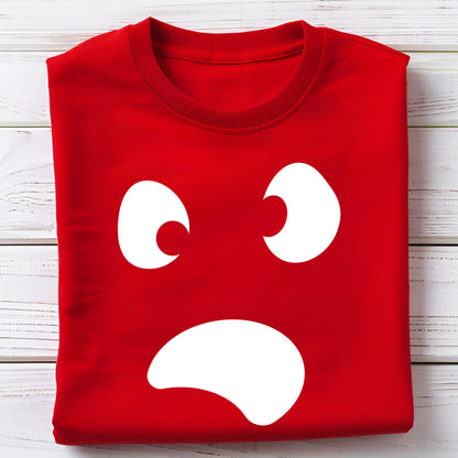 May include: A folded red sweatshirt featuring a white cartoon face design. The design includes two large, curved eyes and a wide, open mouth, creating a surprised or shocked expression. The sweatshirt is on a white wooden surface.