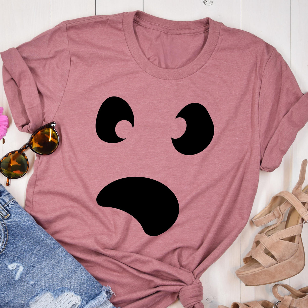May include: Dusty rose t-shirt featuring a large, black, cartoon ghost face. The ghost has two curved eyes and a wide, curved mouth. The shirt is folded and styled with sunglasses and sandals.