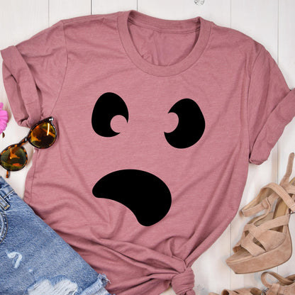 May include: Dusty rose t-shirt featuring a large, black, cartoon ghost face. The ghost has two curved eyes and a wide, curved mouth. The shirt is folded and styled with sunglasses and sandals.