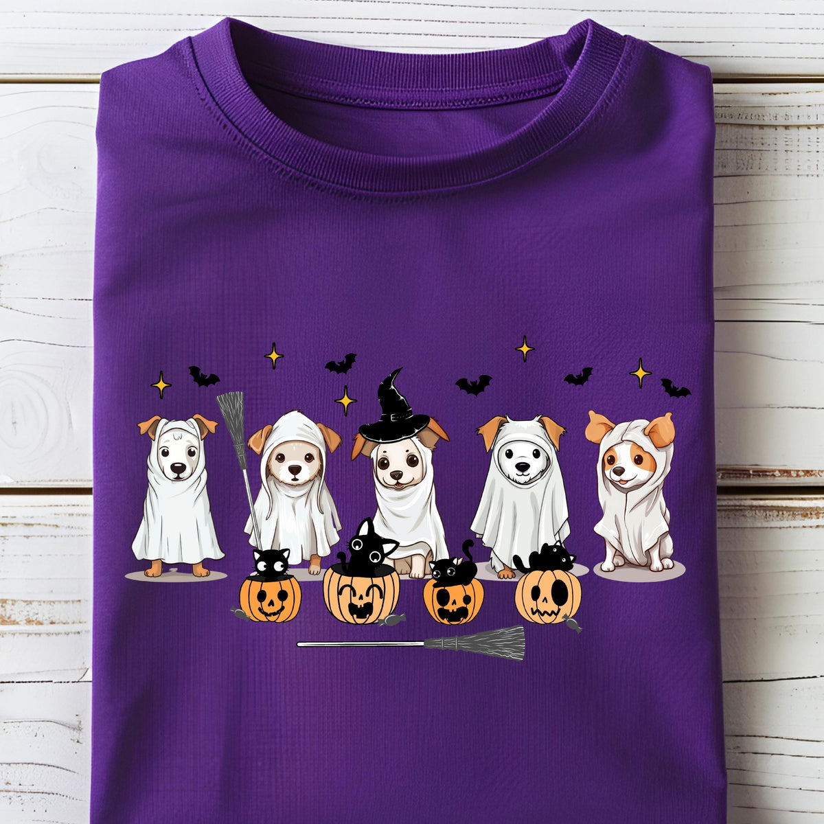May include: A purple t-shirt featuring a graphic print of five dogs wearing white ghost costumes and witch hats, accompanied by four orange jack-o'-lanterns with black cats inside, small black bats, and yellow stars scattered across the design.