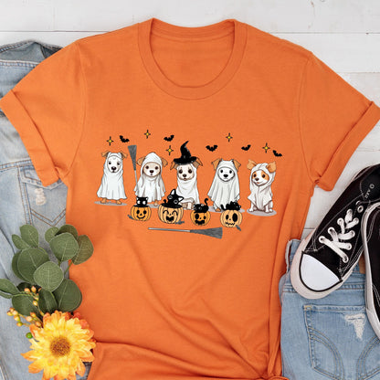 May include: An orange short-sleeved t-shirt featuring a graphic print of five dogs in white ghost costumes, some wearing witch hats, alongside small black cats sitting in carved orange pumpkins, with black bats and yellow stars in the background.