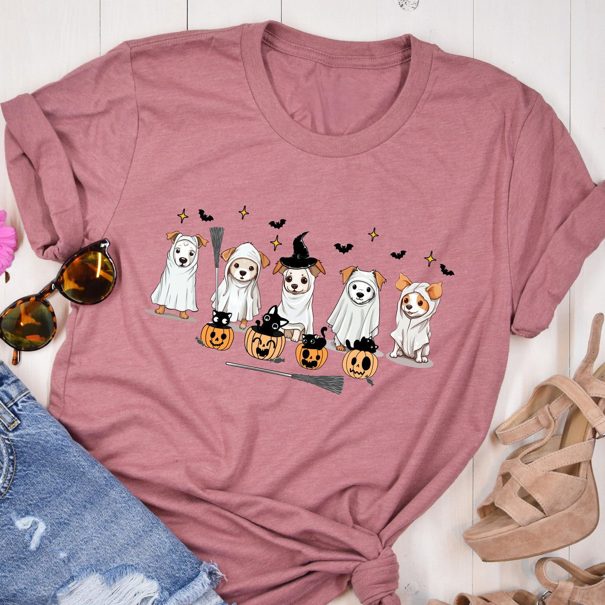 May include: A mauve crew neck t-shirt featuring a graphic print of five dogs dressed in white ghost costumes and witch hats, accompanied by small black cats sitting inside orange jack-o'-lantern pumpkins, black bats, and a broomstick.