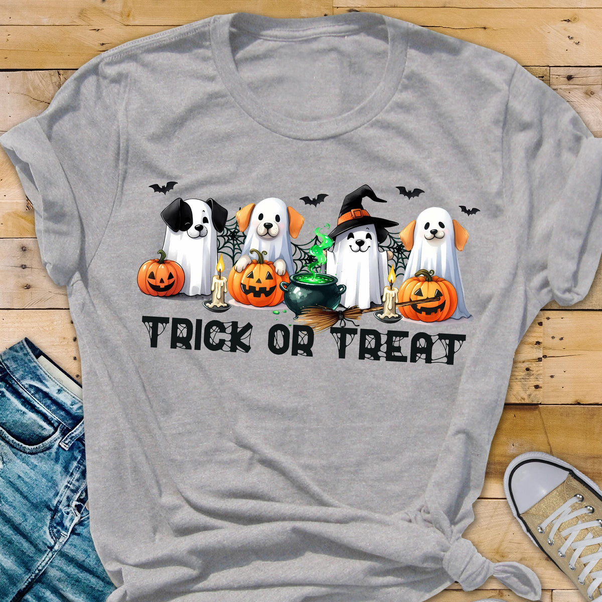 May include: A light gray t-shirt featuring a graphic of four dogs dressed as ghosts for Halloween. The dogs are positioned with pumpkins, a green bubbling cauldron, and candles. The text "TRICK OR TREAT" is printed in black below the dogs.