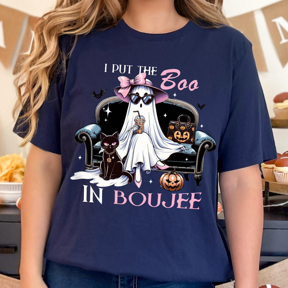 May include: A navy blue t-shirt featuring a graphic of a ghost wearing a pink hat and sunglasses, sitting on a black sofa with a black cat, a pumpkin, and a handbag. The text reads I PUT THE BOO IN BOUJEE in white and pink lettering.