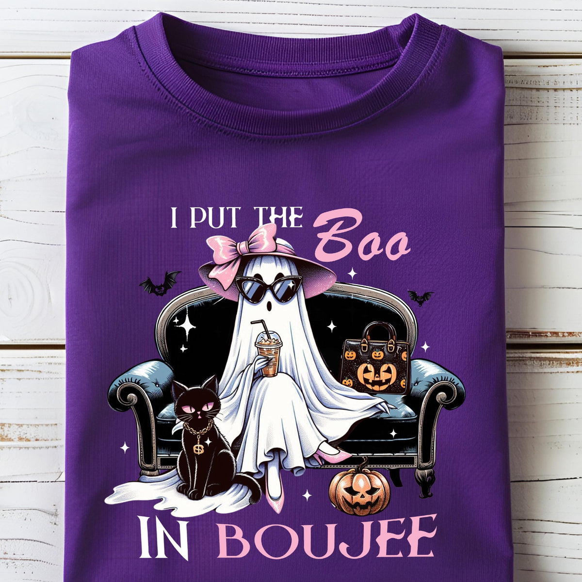 May include: A purple t-shirt featuring a graphic of a white ghost wearing a pink bow, hat, and sunglasses, sitting on a black sofa with a black cat, a pumpkin, and a handbag. The text reads I PUT THE BOO IN BOUJEE with small bats and stars.