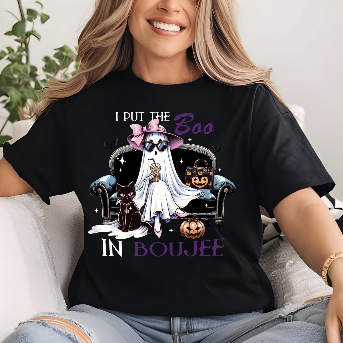 May include: A black t-shirt featuring a graphic of a ghost wearing a pink bow and sunglasses, sitting on a couch with a black cat and a pumpkin-patterned handbag. The text reads I PUT THE BOO IN BOUJEE in white and purple lettering.