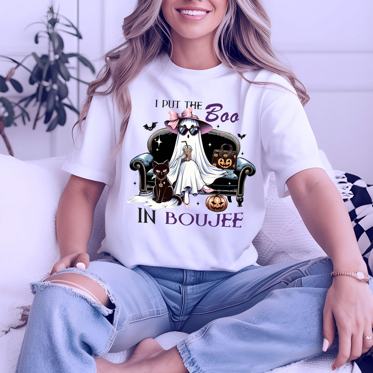 May include: A white t-shirt featuring a graphic of a ghost wearing a pink hat and sunglasses, sitting on a dark sofa with a black cat and a pumpkin. The text reads I PUT THE Boo IN BOUJEE. The design includes small bats and stars in purple and black.