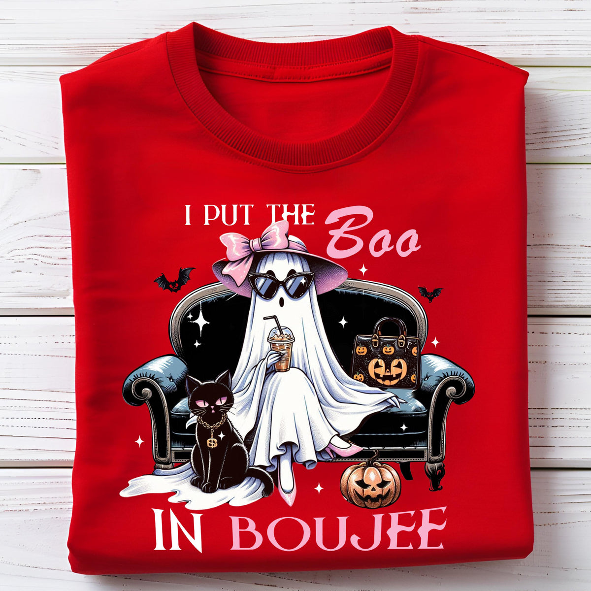 May include: A bright red t-shirt featuring a graphic of a white ghost wearing a pink bow, sunglasses, and holding a coffee cup while sitting on a black sofa next to a black cat and a pumpkin, with the text I PUT THE BOO IN BOUJEE printed.