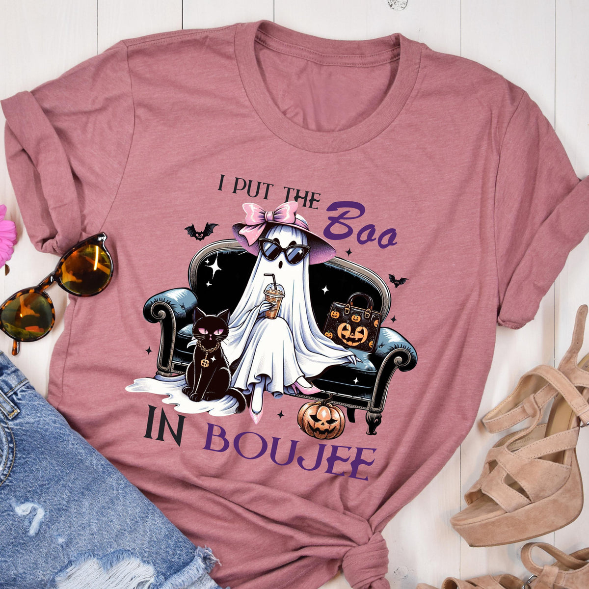 May include: A mauve short-sleeve t-shirt featuring a graphic of a ghost wearing a pink bow and sunglasses, sitting on a black sofa with a black cat and a pumpkin-patterned handbag. The text reads I PUT THE BOO IN BOUJEE with small bats.