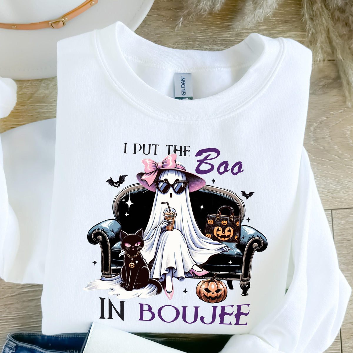 May include: A white long-sleeve sweatshirt featuring a graphic of a ghost wearing a pink bow, sunglasses, and holding a drink while sitting on a black sofa. A black cat, a pumpkin-patterned handbag, and a jack-o'-lantern are present with the text I PUT THE BOO IN BOUJEE.