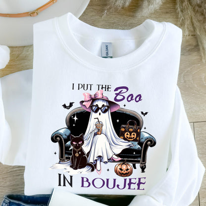 May include: A white long-sleeve sweatshirt featuring a graphic of a ghost wearing a pink bow, sunglasses, and holding a drink while sitting on a black sofa. A black cat, a pumpkin-patterned handbag, and a jack-o'-lantern are present with the text I PUT THE BOO IN BOUJEE.