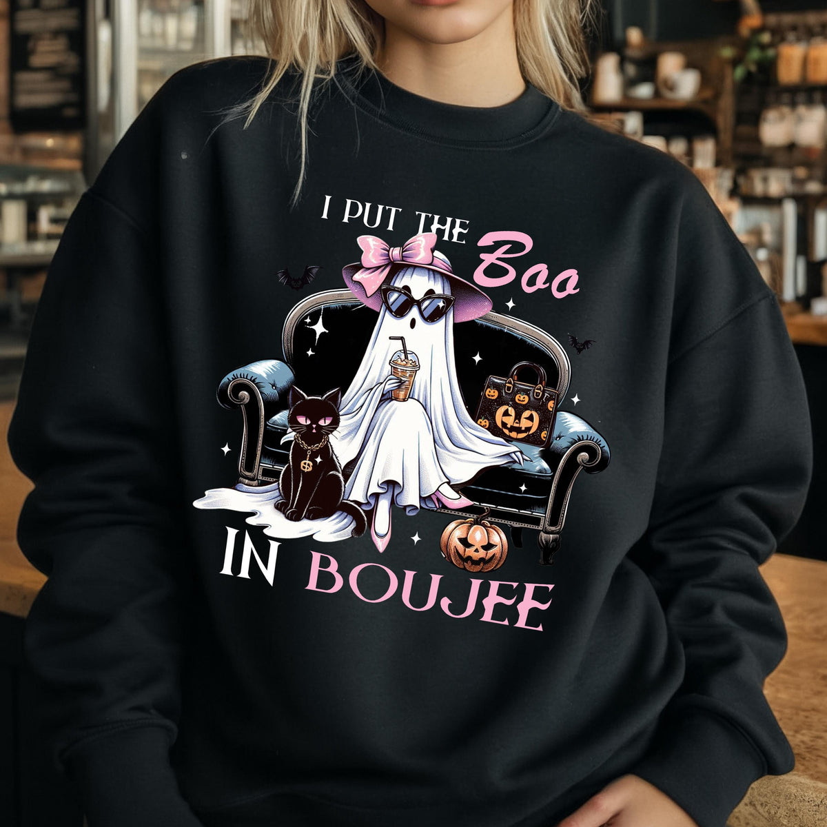May include: A black crewneck sweatshirt featuring a graphic of a ghost wearing a pink bow, hat, and sunglasses, sitting on a sofa with a black cat, a pumpkin, and a handbag. The text reads I PUT THE BOO IN BOUJEE in white and pink lettering.