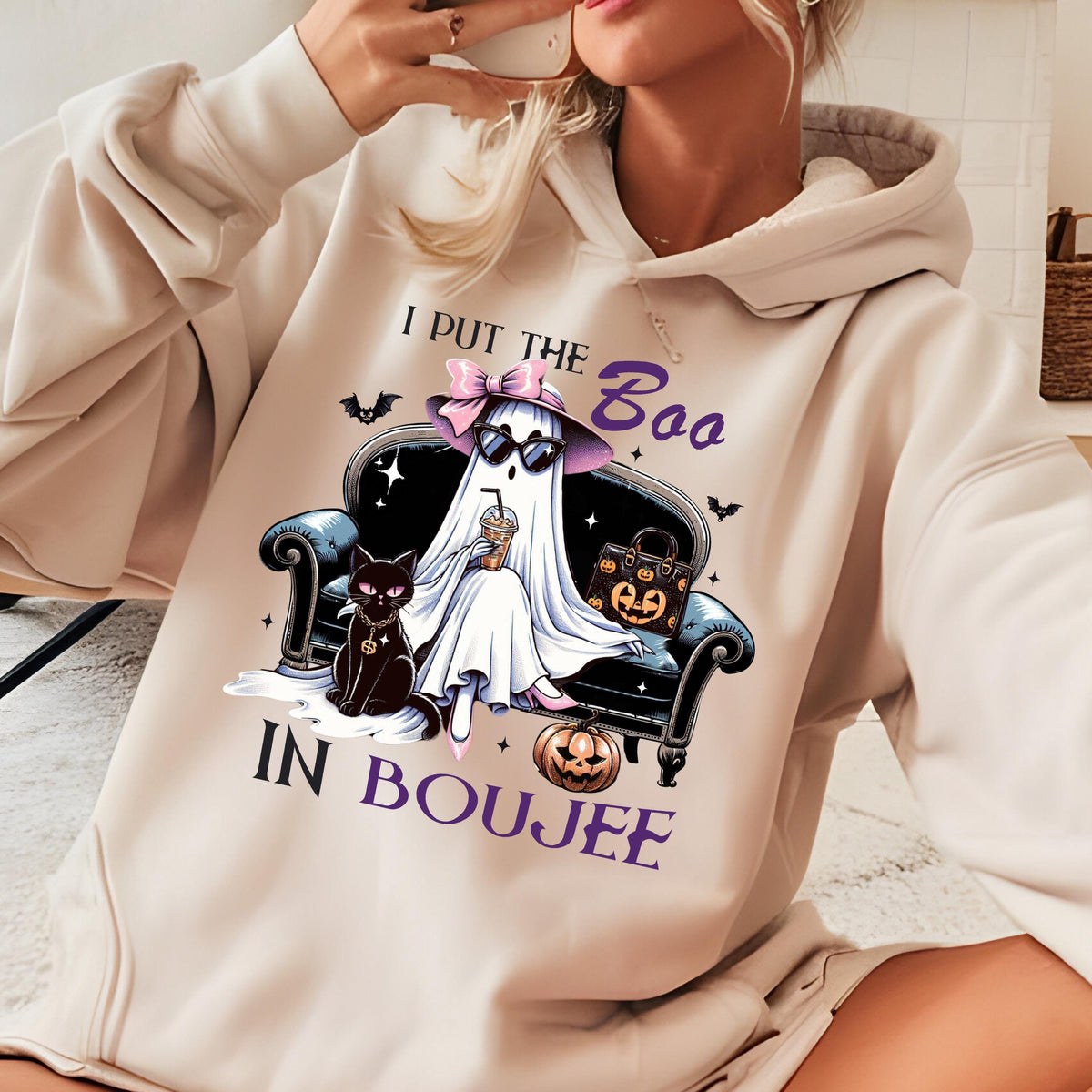 May include: A beige hooded sweatshirt featuring a graphic of a ghost wearing a pink bow, sunglasses, and holding a drink while sitting on a black sofa next to a black cat and a pumpkin. The text reads I PUT THE BOO IN BOUJEE in purple lettering.