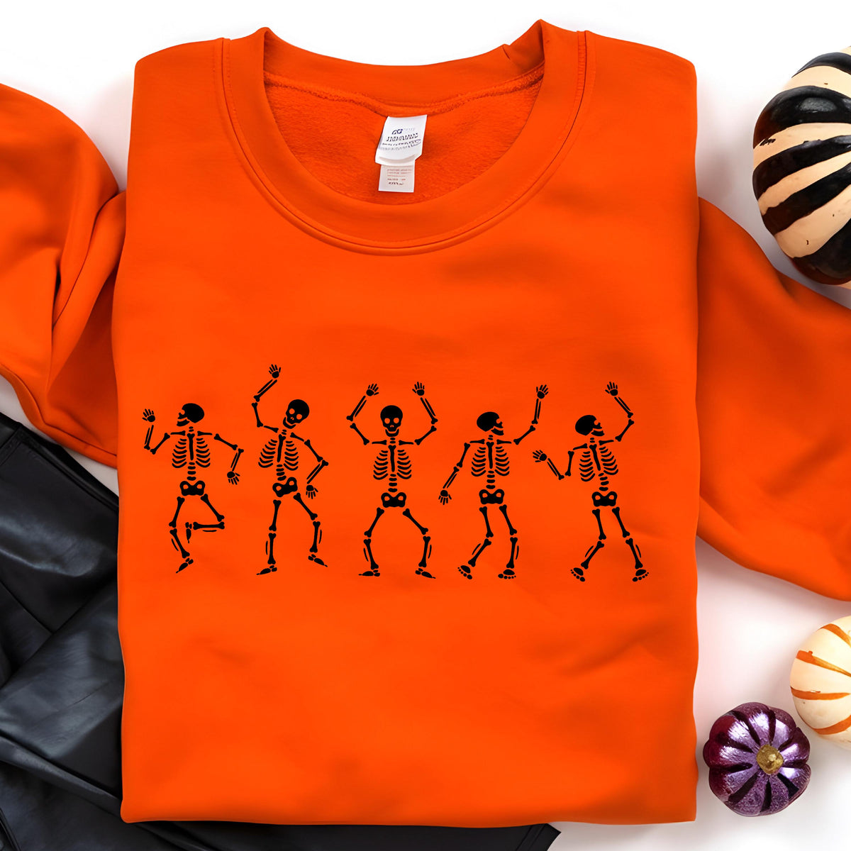 May include: An orange sweatshirt featuring a black graphic of five dancing skeletons. The skeletons are in various poses with their arms raised. The sweatshirt is a solid orange color, perfect for Halloween.