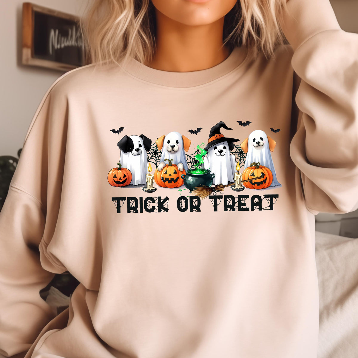 May include: Beige sweatshirt featuring a Halloween-themed graphic. The design includes four dogs dressed as ghosts, pumpkins, bats, candles, and a witch's cauldron. The words "TRICK OR TREAT" are printed in black block letters below the graphic.