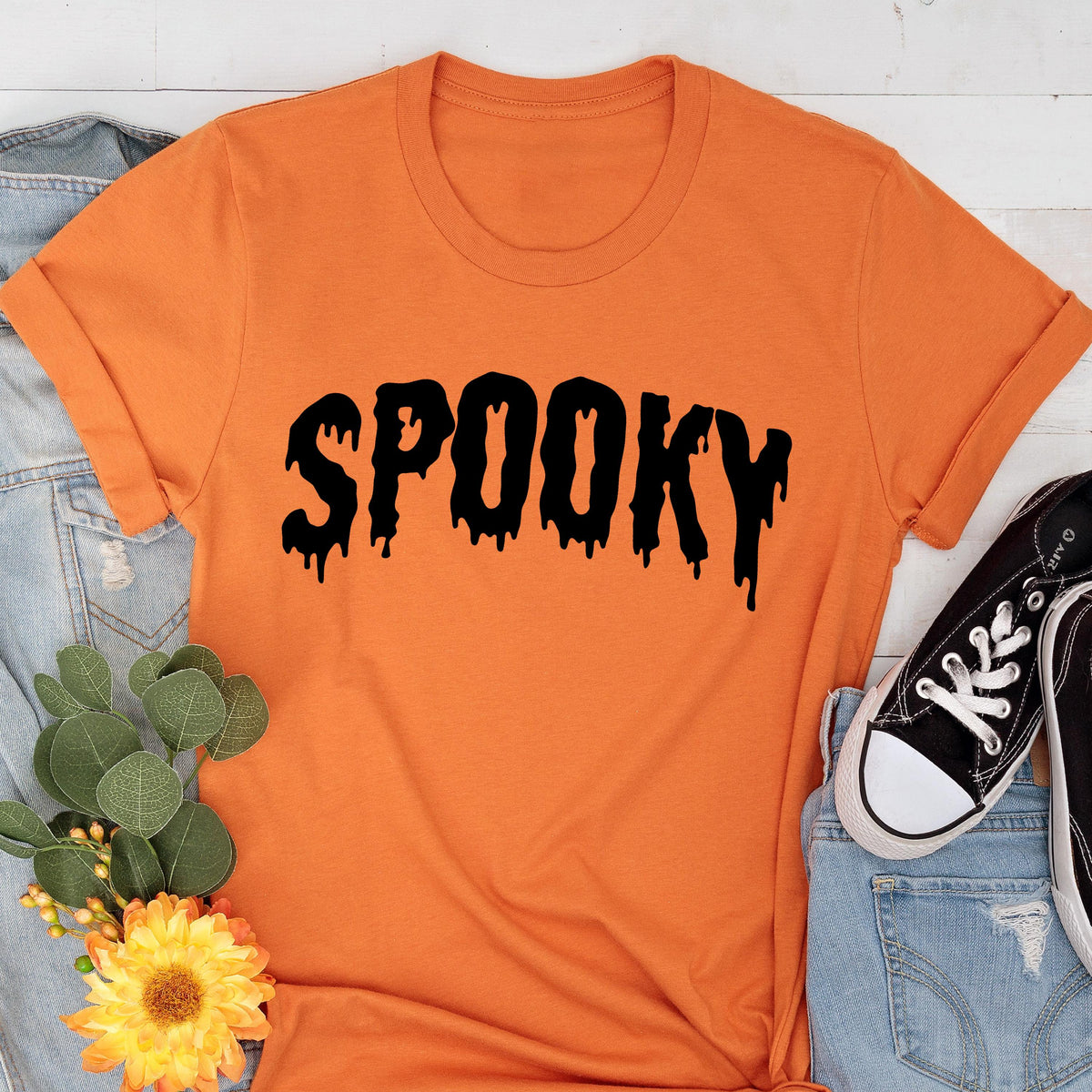May include: Orange t-shirt with the word "SPOOKY" in black, dripping font. The shirt is displayed with a pair of black sneakers, blue jeans, and a floral arrangement, creating a Halloween theme.