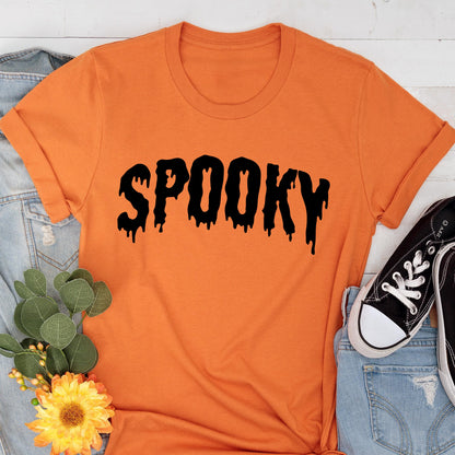 May include: Orange t-shirt with the word "SPOOKY" in black, dripping font. The shirt is displayed with a pair of black sneakers, blue jeans, and a floral arrangement, creating a Halloween theme.