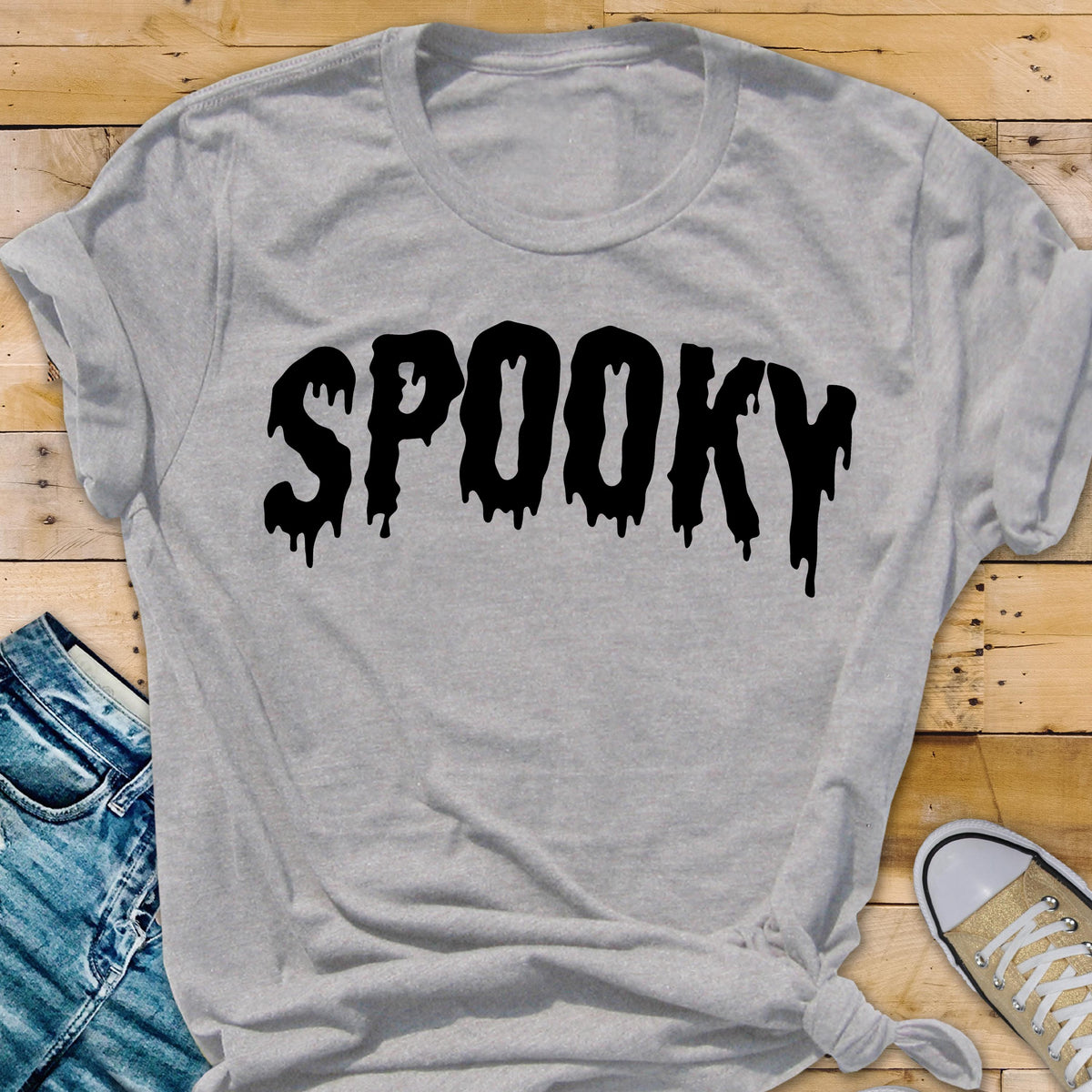 May include: A light gray t-shirt with the word "SPOOKY" in large, black, dripping letters. The shirt is a casual style, with rolled-up sleeves and a knotted hem. The design is perfect for Halloween.