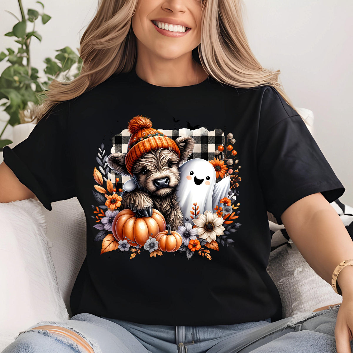 May include: A black t-shirt featuring a graphic print of a fluffy highland cow wearing an orange knit beanie, accompanied by a small white ghost, orange pumpkins, and autumn flowers set against a black and white buffalo plaid background.