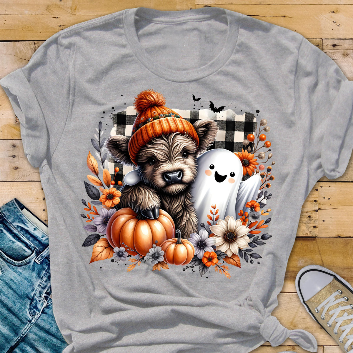 May include: A light gray t-shirt featuring a graphic of a fluffy highland cow wearing an orange knit hat, sitting next to a white ghost. The design includes orange pumpkins, autumn flowers, and a black and white buffalo plaid background.