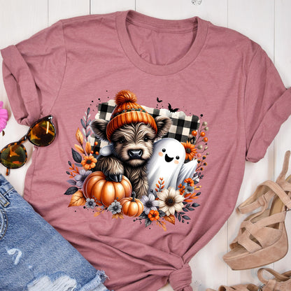 May include: A mauve t-shirt featuring a graphic print of a fluffy highland cow wearing an orange knit hat, sitting next to a white ghost. The design includes orange pumpkins, autumn flowers, and a black and white buffalo plaid square background.