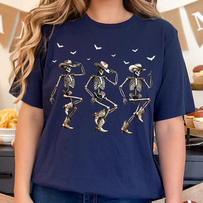 May include: Navy blue t-shirt featuring a graphic of three dancing skeletons wearing cowboy hats and boots. White bats are scattered above the skeletons. The skeletons are in various dance poses, creating a fun and festive design.