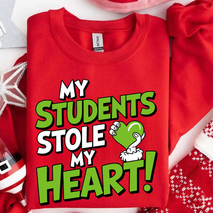 May include: A red crewneck sweatshirt with the text "MY STUDENTS STOLE MY HEART!" in white and green. The design includes a green heart held by a hand. The sweatshirt is a festive item, possibly for a teacher.