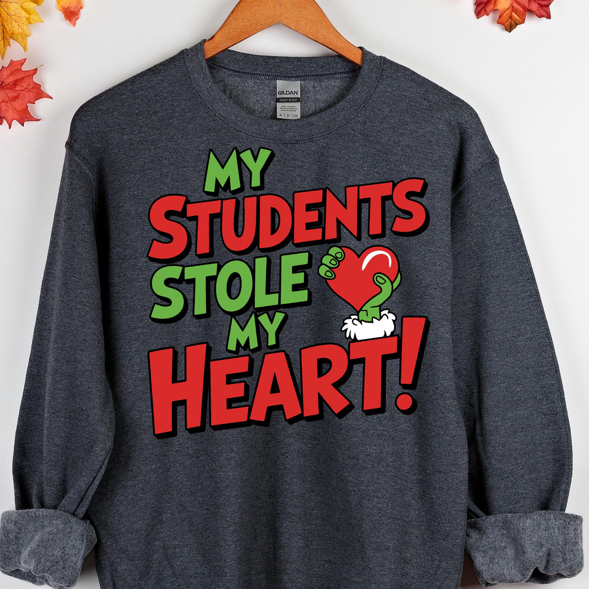 May include: Dark gray sweatshirt with the text "MY STUDENTS STOLE MY HEART!" in green and red. The design includes a red heart held by a green hand. The sweatshirt is hanging on a wooden hanger.